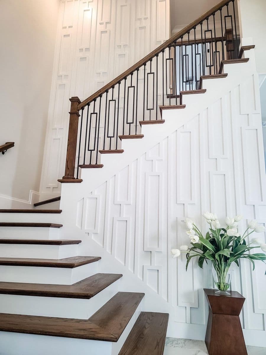 White accent wall with geometric raised panels beside a wooden staircase in a modern home, perfect for wallpaper installation