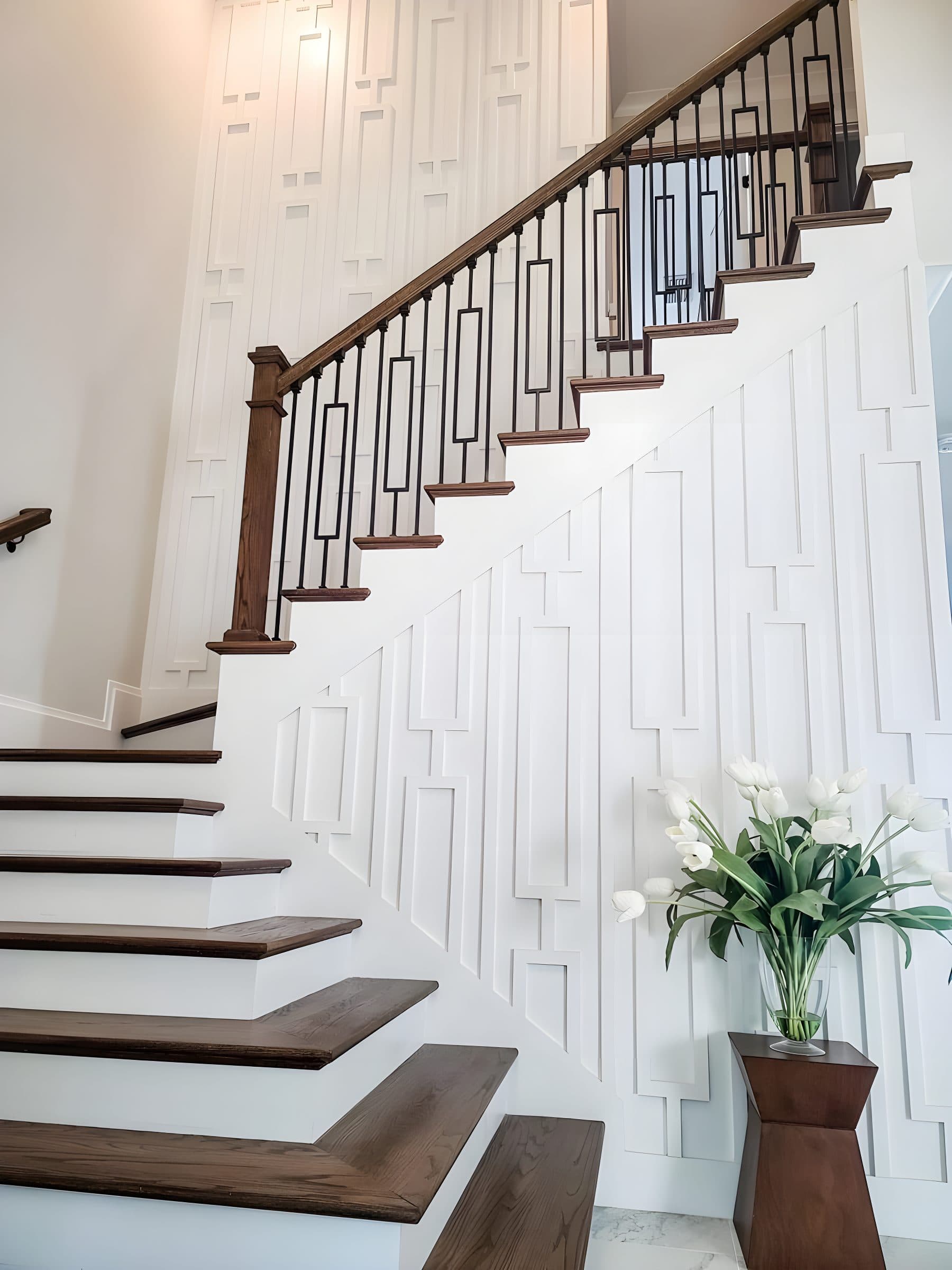 White accent wall with geometric raised panels beside a wooden staircase in a modern home, perfect for wallpaper installation